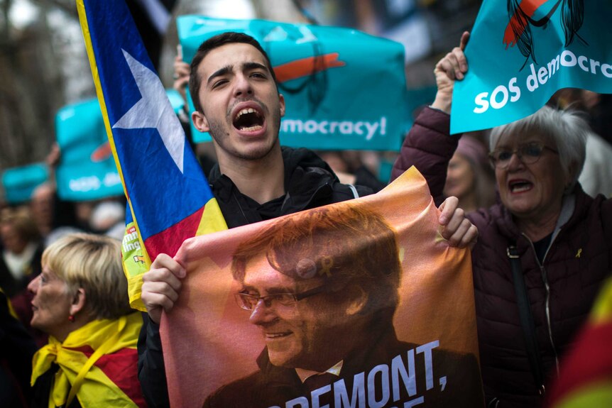 A demonstrator holds a Puigdemont poster and yells, he is also carrying a Catalan flag.