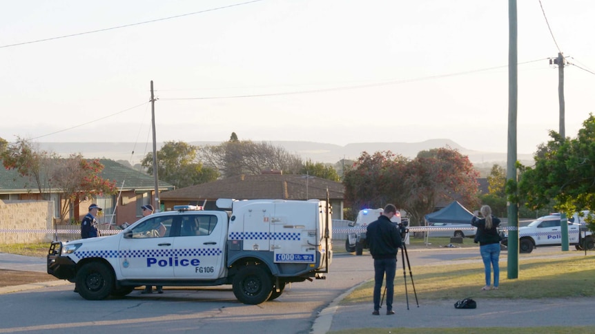 A police utility sits parked across a street which is cordoned off with police tape with two more police cars in the background.