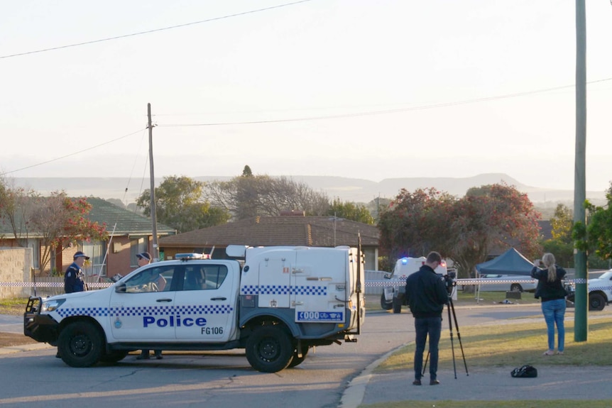 A police utility sits parked across a street which is cordoned off with police tape with two more police cars in the background.