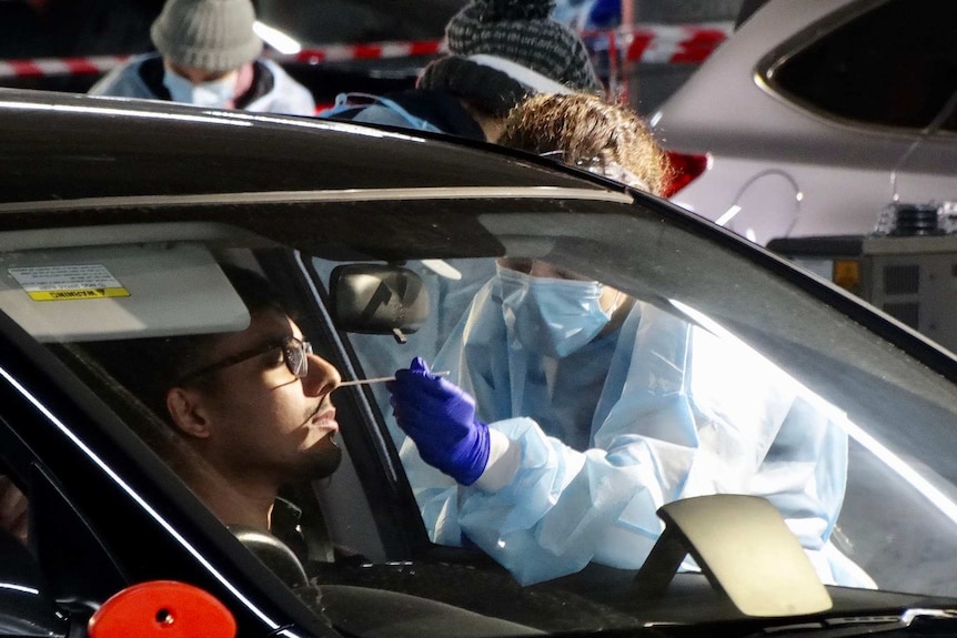 A man sits in a car and gets a coronavirus test by a woman in full personal protective gear.