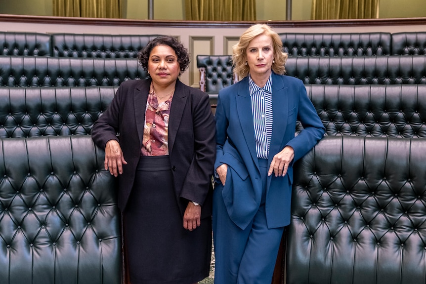 Deborah Mailman and Rachel Griffiths stand side-byside in a parliamentary chamber. They lean on the leather chairs