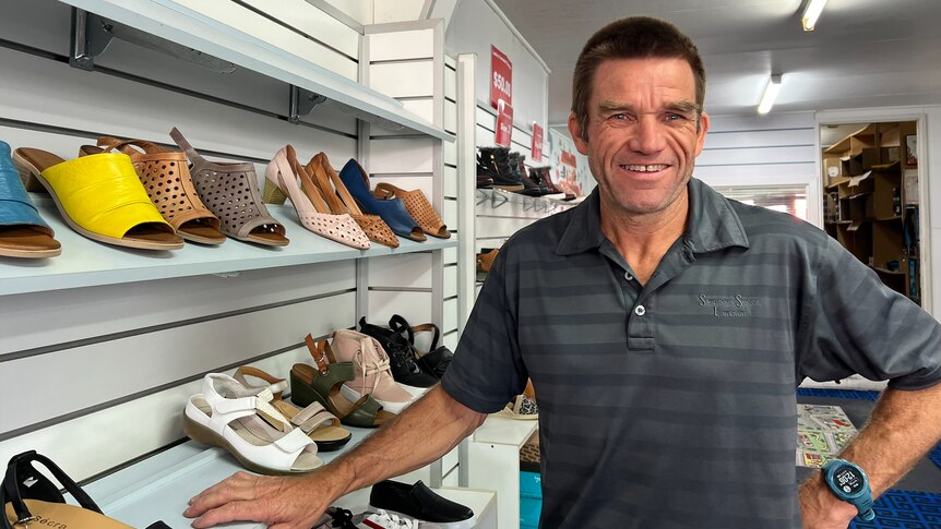 A man wearing a grey shirt and a big smile stand besides a shoe rack in a shoe store