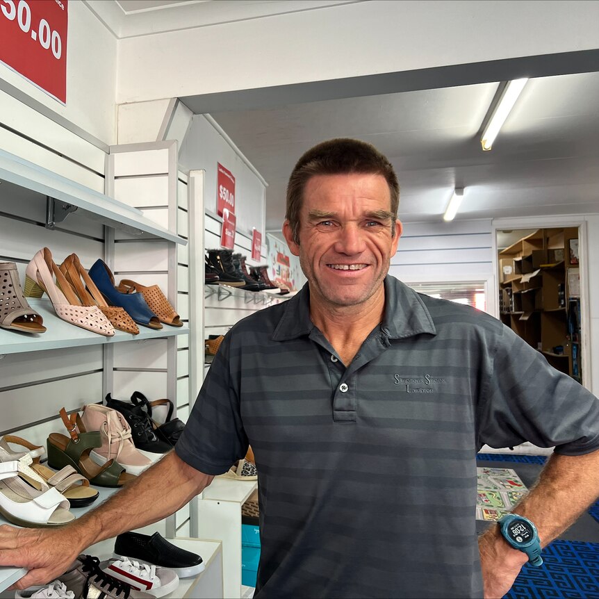 A man wearing a grey shirt and a big smile stand besides a shoe rack in a shoe store