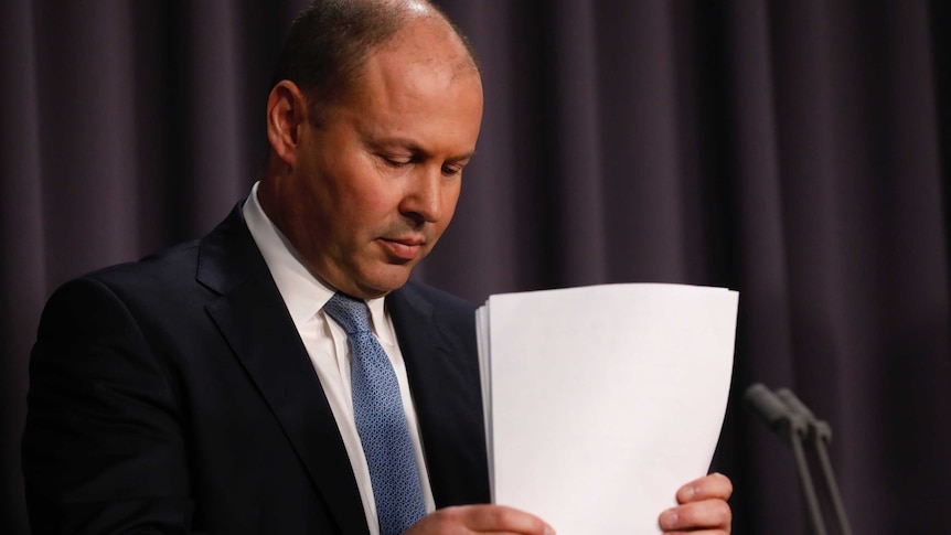Josh Frydenberg looks down while shuffling papers at a lectern