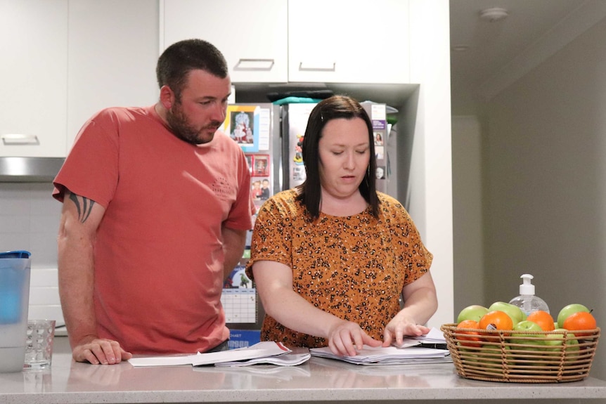 A 30-something couple in casual clothes stand and look over documents on a beige kitchen counter.