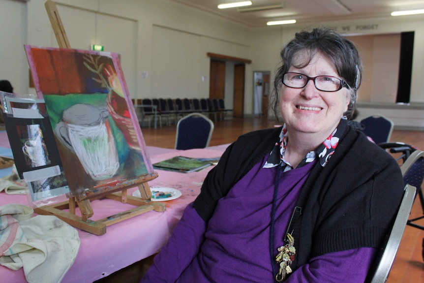 A woman with dark hair wearing a purple jumper, sitting next to a small easel on a table.