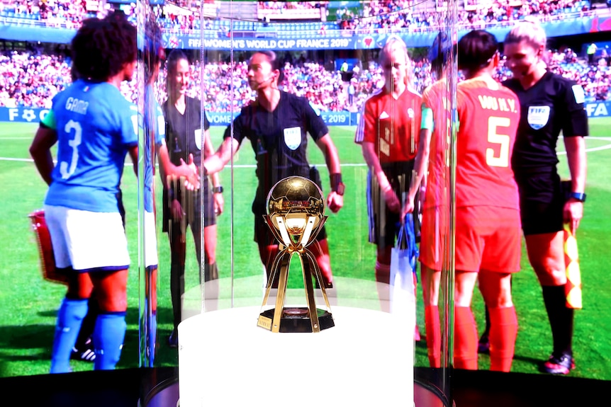 A trophy inside a glass case sits in front of a film playing highlights of a women's soccer game