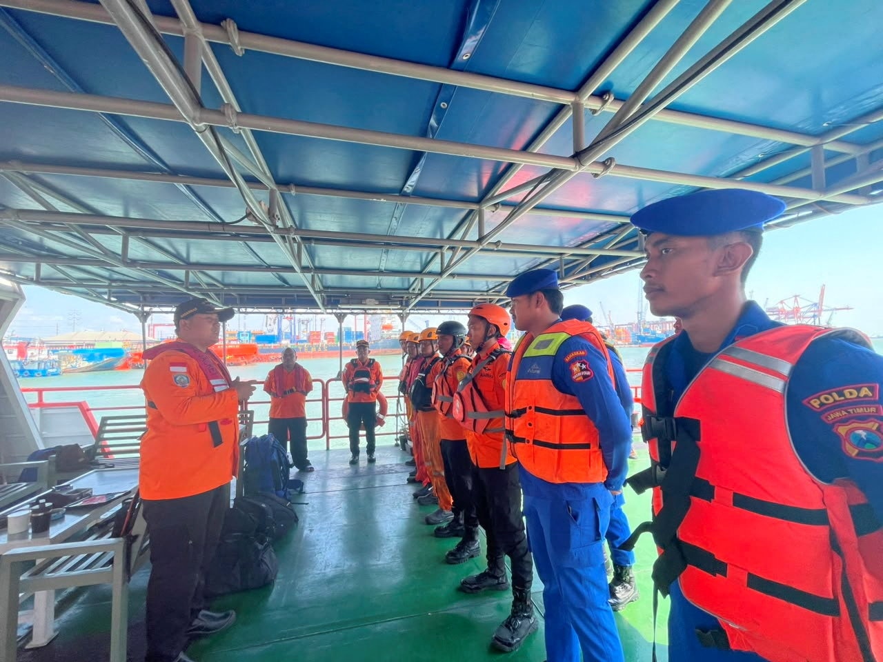 A group of men in orange life jackets are being briefed on a ship