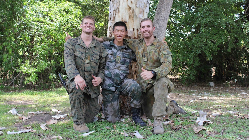 Three men in army and US marines uniforms kneel before a tree with their arms around each other.
