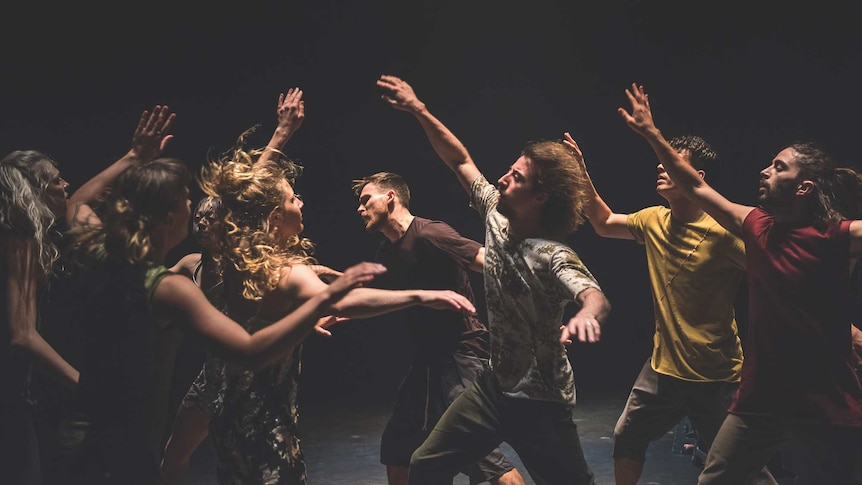 Two groups of dancers on a black stage march towards each other with their arms raised.