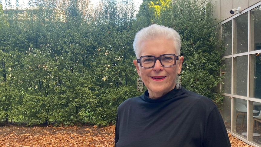 Woman with white hair and glasses smiles, in front of a tall hedge