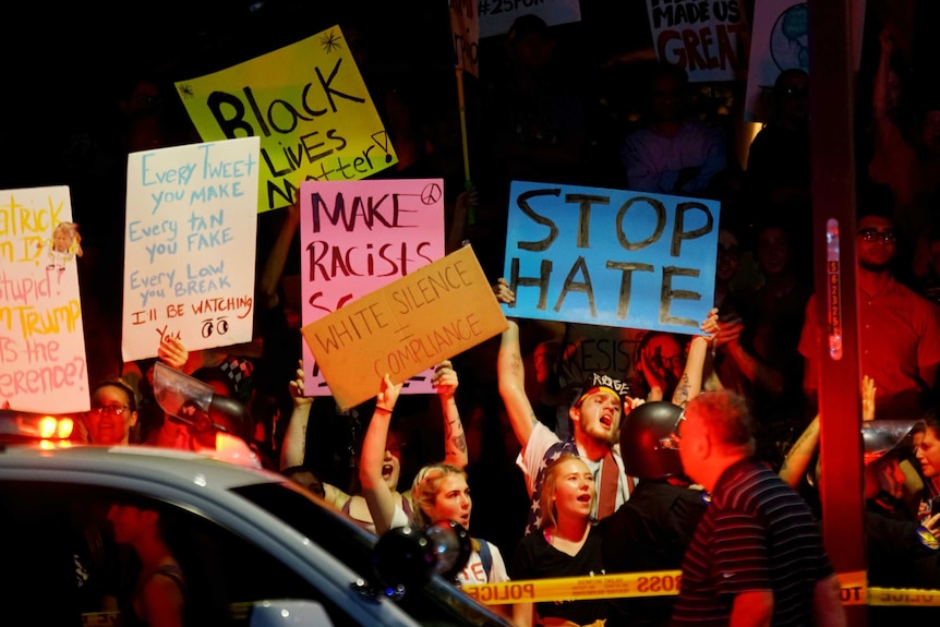 A crowd of people hold anti-Trump signs in the evening. They are behind yellow police tape