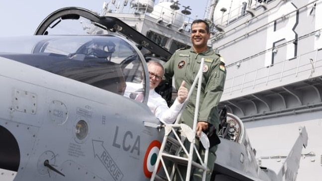 A man in a white shirt gives a thumb-up while leaning out of a fighter jet.
