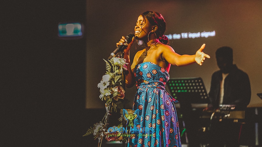 Elsy Wameyo stands on stage with her arm held out to her side singing into a microphone