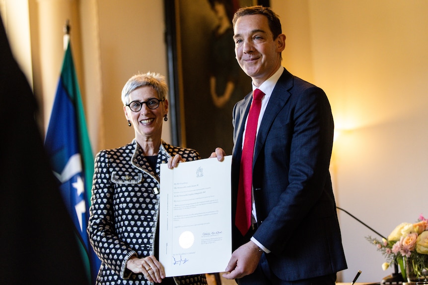 A man and woman in formal attire smile and hold up a document.