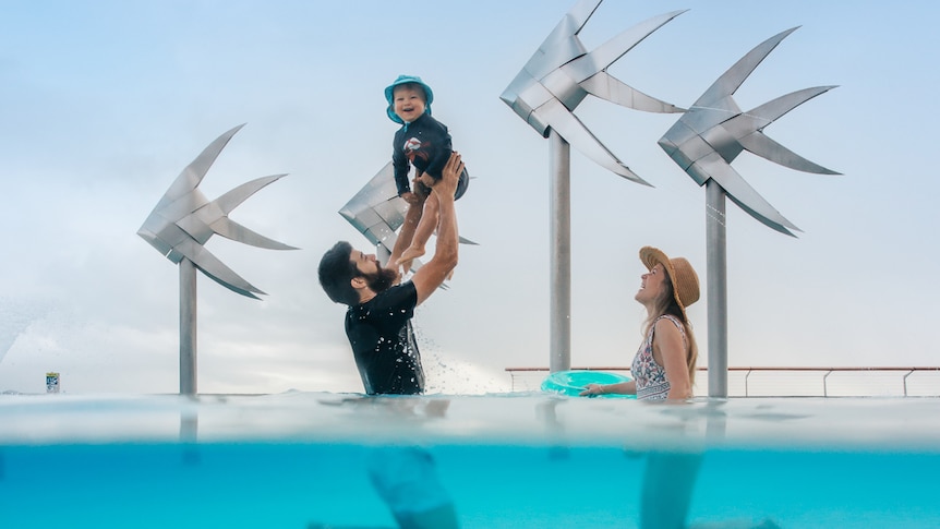 A woman stands next to a man holding a smiling toddler above his head in a large pool, with metal fish sculptures behind them.