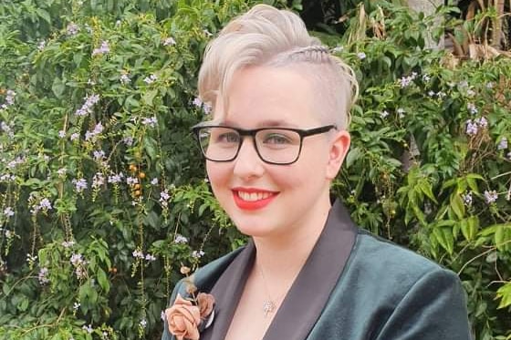 A person, who identifies as non-binary, stand in a green velvet tux in front of a flowering bush.