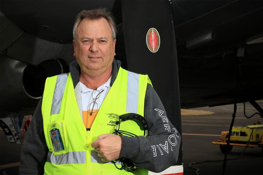 An older man in high-vis stands holding aviation headphones in front of a plane.