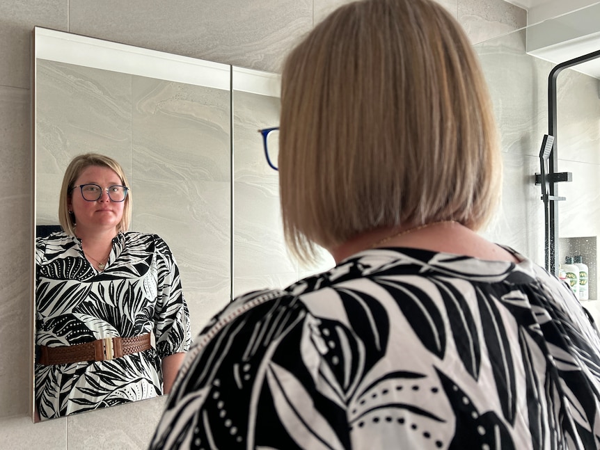 A woman with eczema and glasses looks at herself in the mirror.