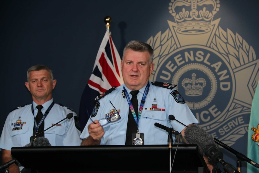 An elderly police officer in uniform holds his glasses at podium as a colleague listens next to him.