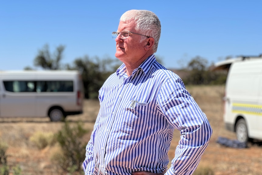 A man with grey hair wearing a striped shirt stares ahead with scrubland and police vans in the background