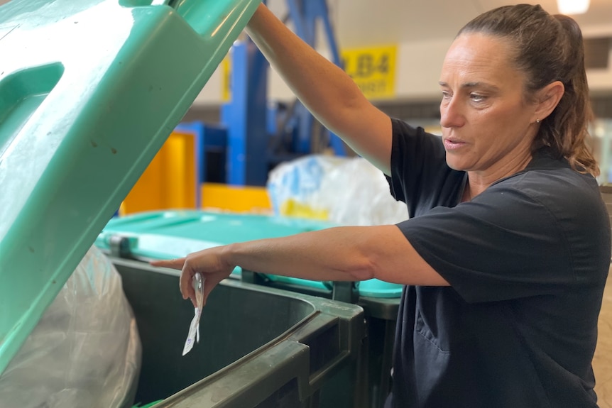 A woman inspects a bin by lifting a baby blue lid and pointing towards a bag of plastic waste