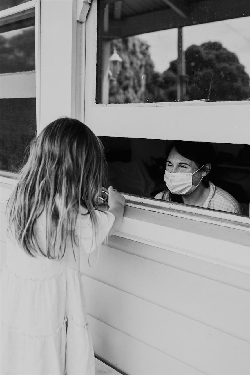 Black and white image of a small girl looking through a partially-opened window at a masked woman inside.