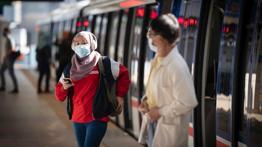 A woman wearing a hijab, headphones, face mask and a man wearing a mask come out of public public transport.