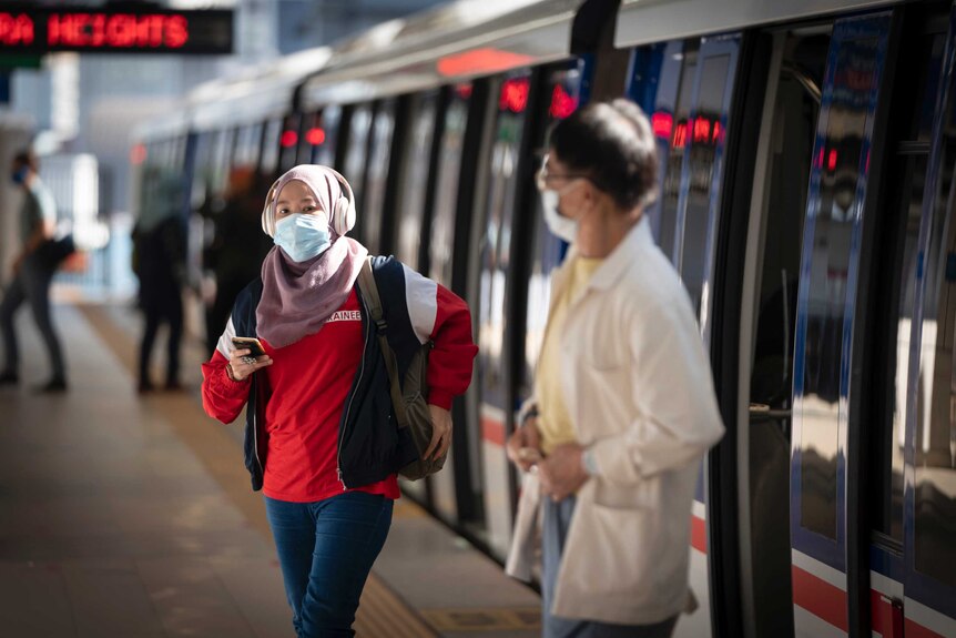 A woman wearing a hijab, headphones, face mask and a man wearing a mask come out of public public transport.