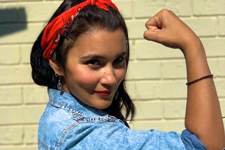 A woman in a blue shirt and red bandana flexes her arm and looks at the camera