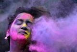 Closeup of a woman's face covered in coloured powders, on the right side a cloud of purple powder.