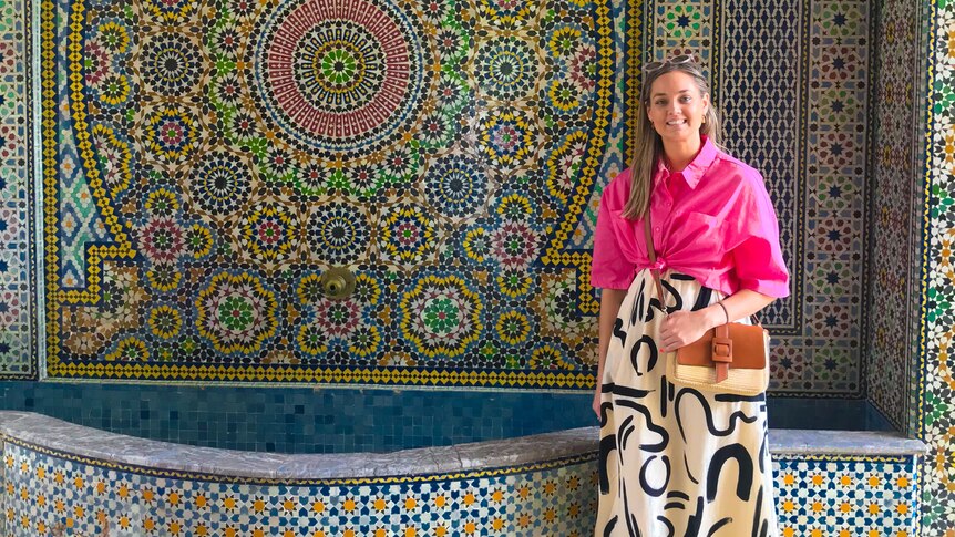 Eliza wears a pink shirt and white and black skirt and smiles in front of a Moroccan tiled fountain