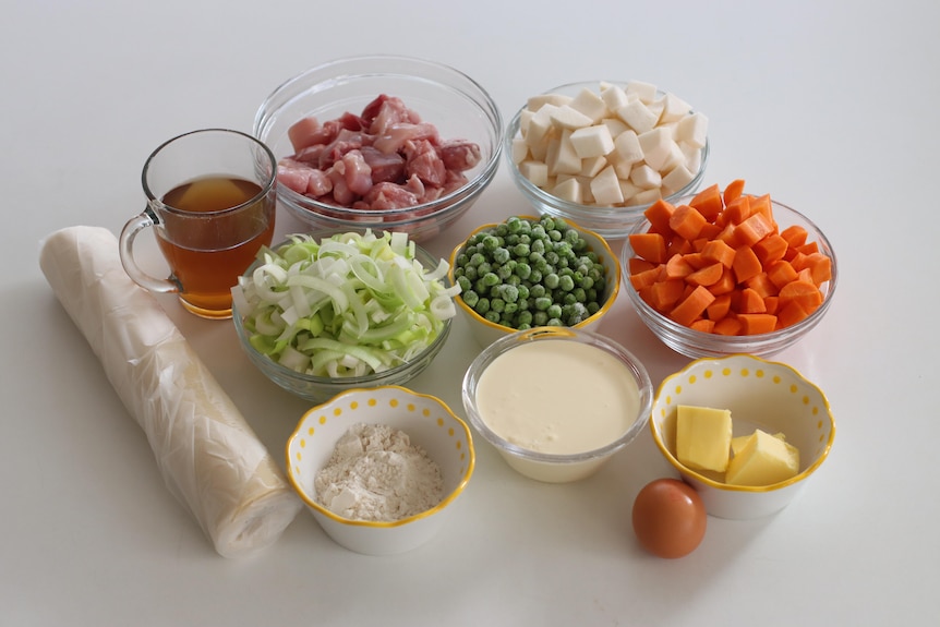 A variety of ingredients, including chicken, peas, carrot and butter, sitting in individual bowls on a bench.