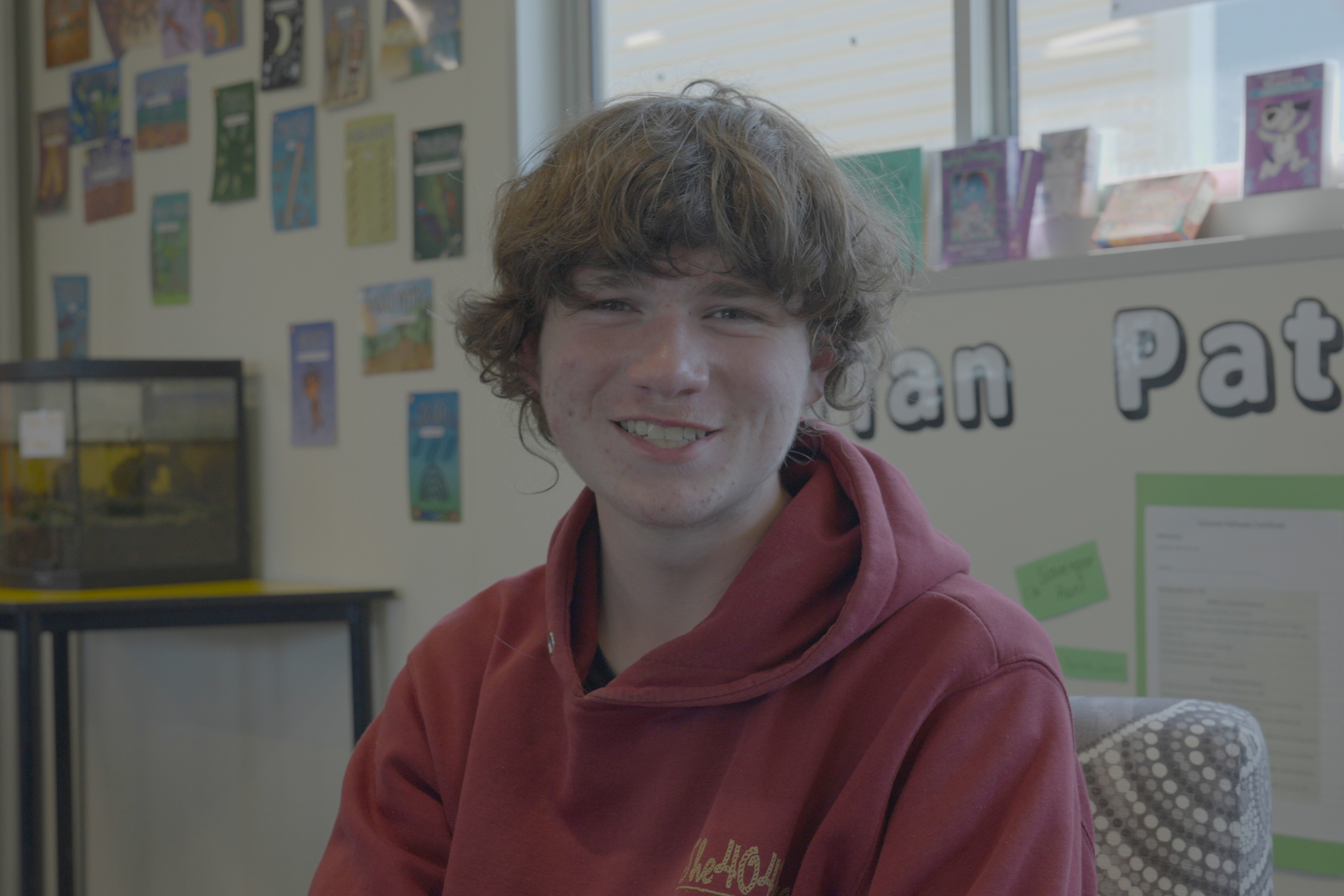 Um adolescente sorri. Ele usa um moletom vermelho e tem cabelo castanho de comprimento médio.