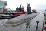 A surfaced nuclear submarine in dock next to a tug boat. 