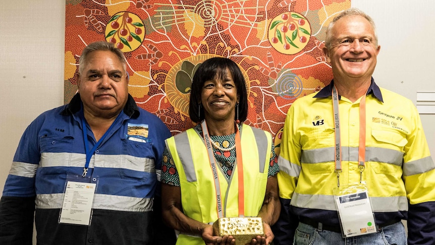 A woman and two men, the woman holding a gold bar in front of an indigenous artwork