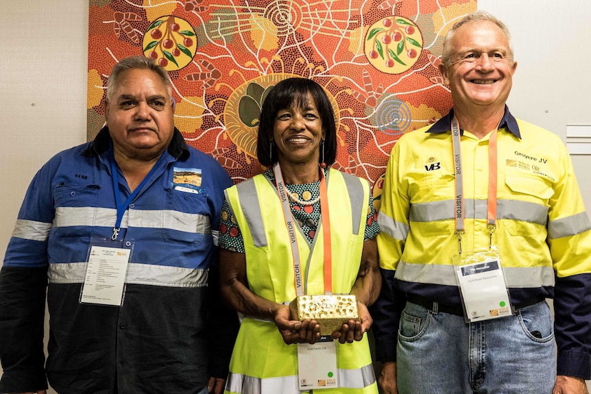 A woman and two men, the woman holding a gold bar in front of an indigenous artwork