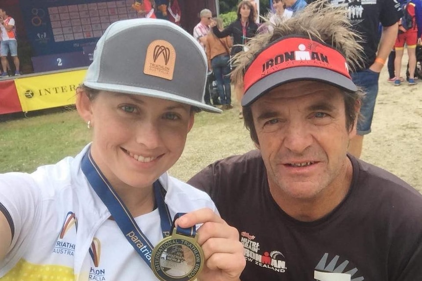 A man and a woman smile at the camera, the woman holding a gold medal.