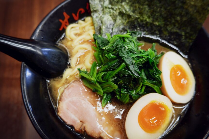 Picture of ramen in a black soup bowl consisting of noodles, two egg halves and green vegetables decorated on top