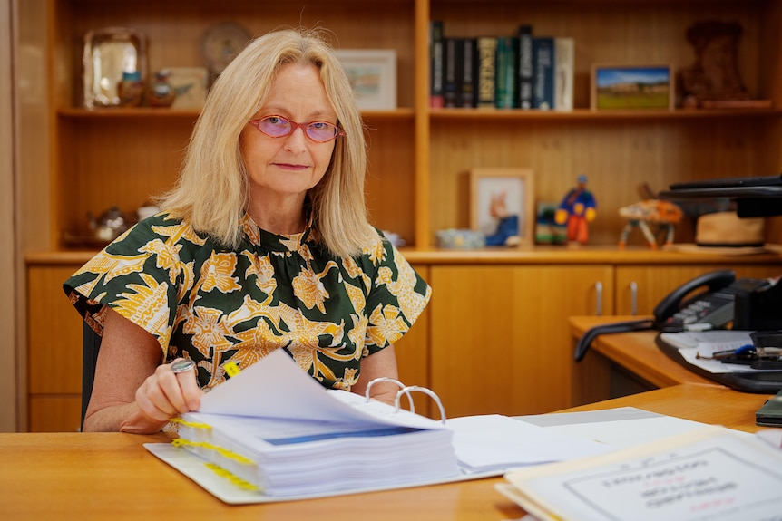 A woman sitting at a desk inside an office and turning pages in a large file.