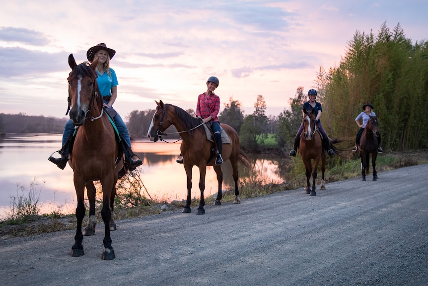 Four riders on horseback standing in a row on a trail along the banks of a river.