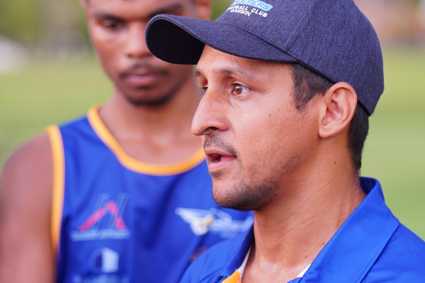 A man wearing a cap talks while a young man in a football jersey stands in the background. 