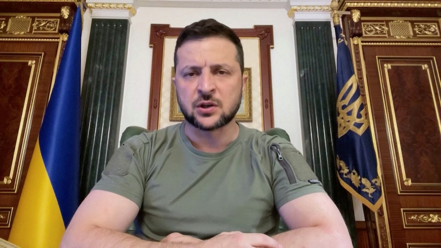 A man with short dark hair wearing combat fatigues speaks to the camera while seated in front of a Ukrainian flag in an office.