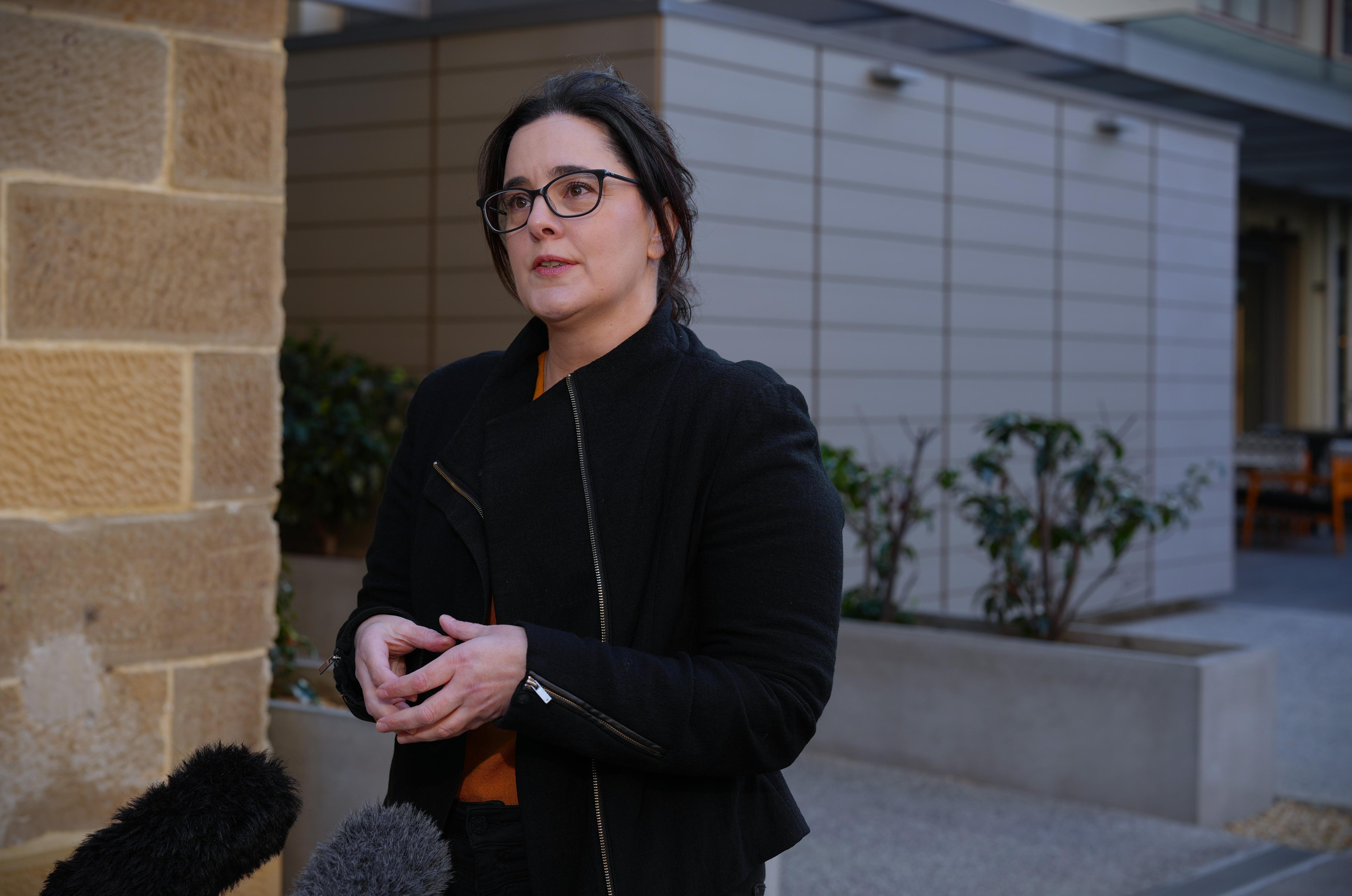 Una mujer con gafas y cabello oscuro, vestida con una chaqueta oscura, habla ante micrófonos frente a un edificio de ladrillo.