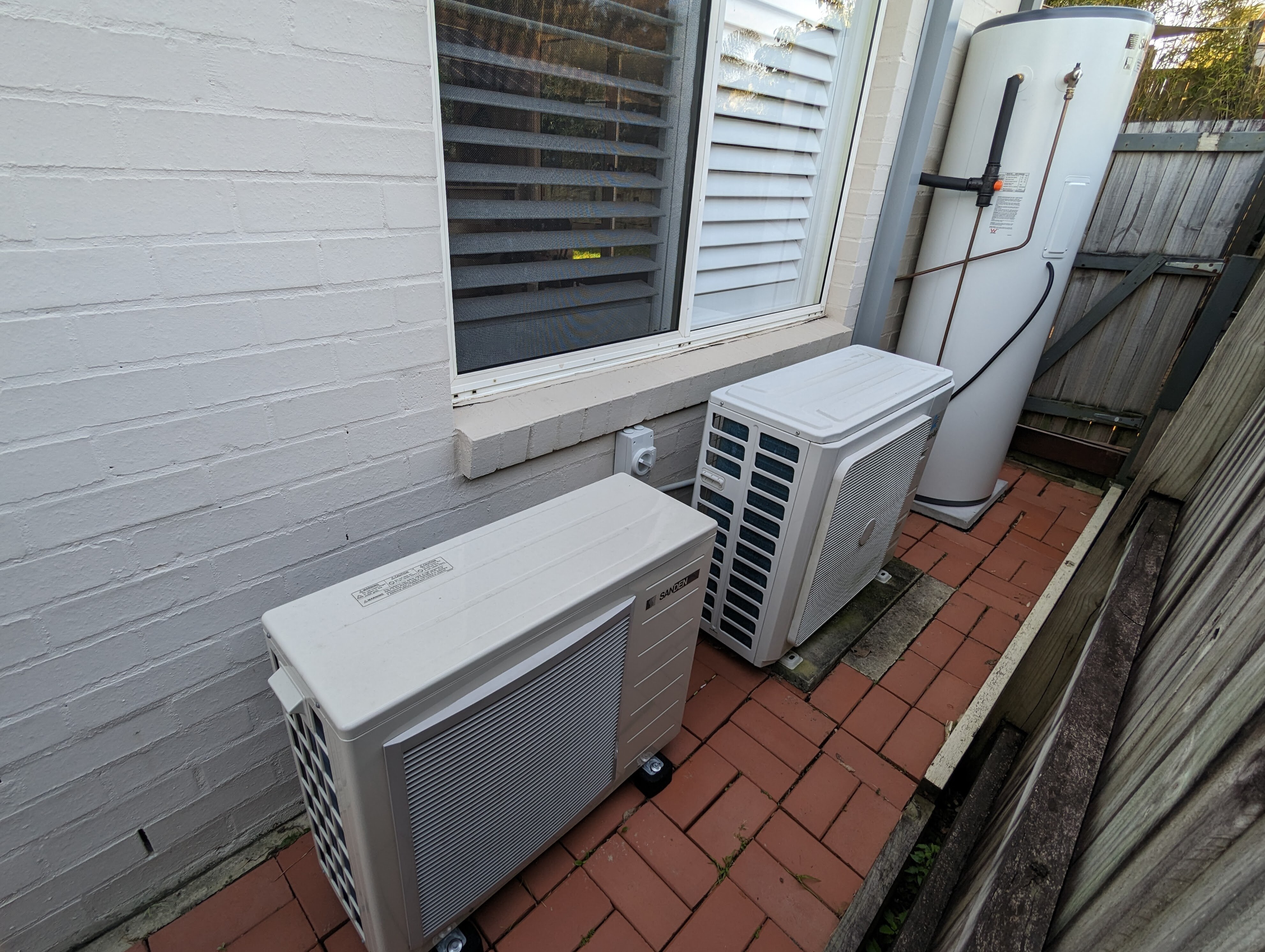 A white heat pump hot water system positioned on pavers outside a white brick home, beside a hot water system.