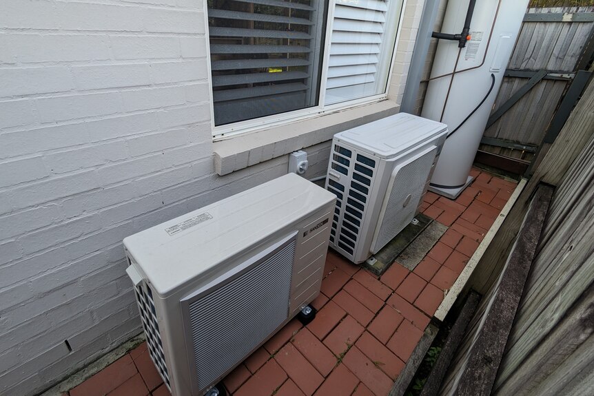 A white heat pump hot water system positioned on pavers outside a white brick home, beside a hot water system.