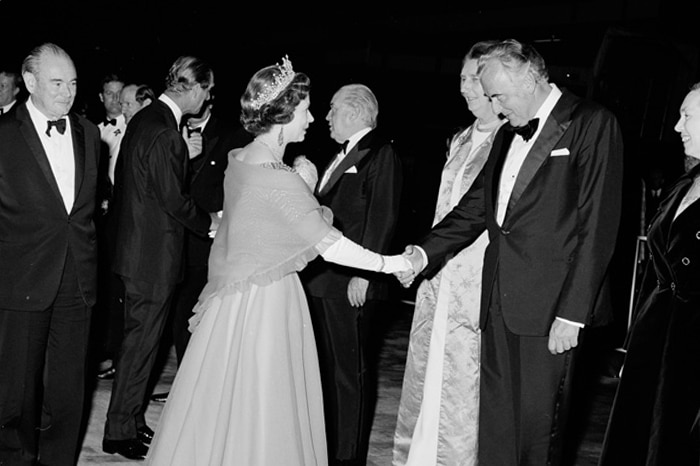 A woman in royal garb shakes the hand of a reverent man in suit and bowtie.