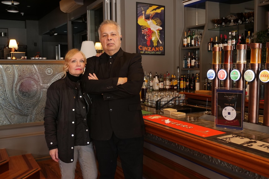 A man wearing black folding his arms alongside a woman wearing black with blonde hair standing his left in a bar setting.