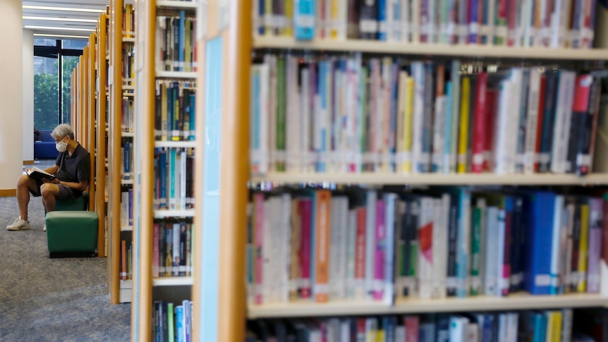 A man sits with books on shelves blurred in foreground.