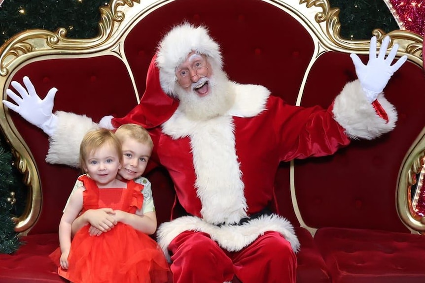 A young girl in a red dress being held by her brother next to Santa.